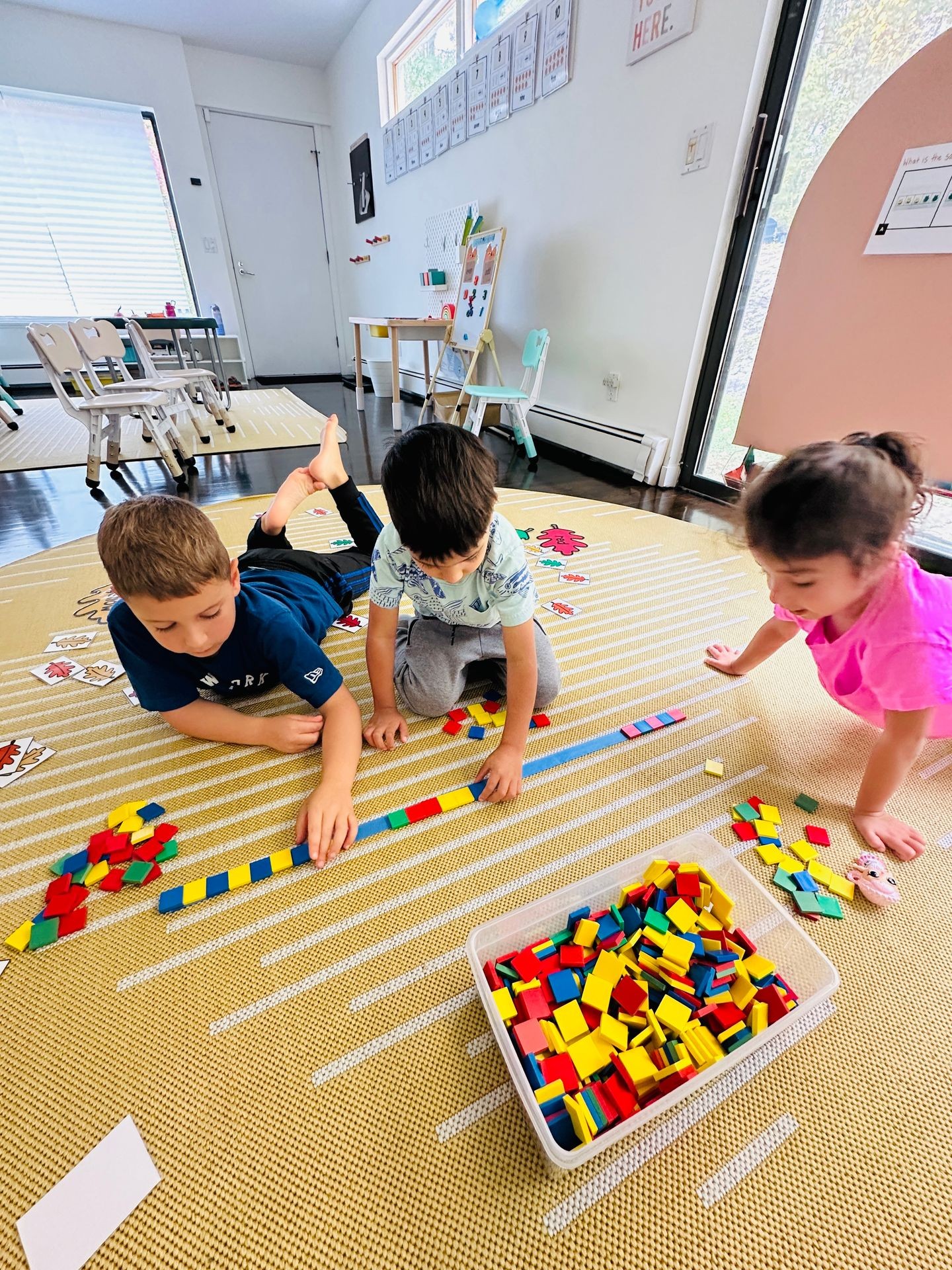 Children sitting on a mat playing with colorful geometric blocks in a classroom setting.