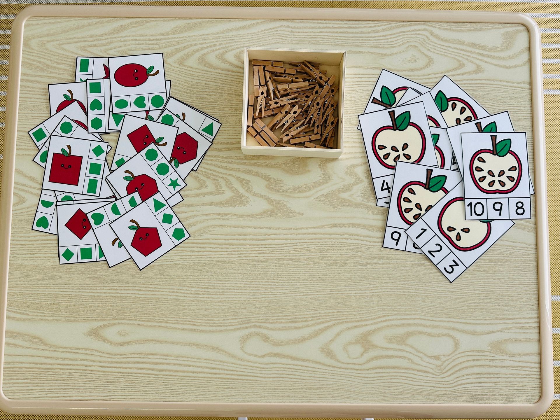Educational cards with apples and shapes on a table, alongside a box of clothespins.