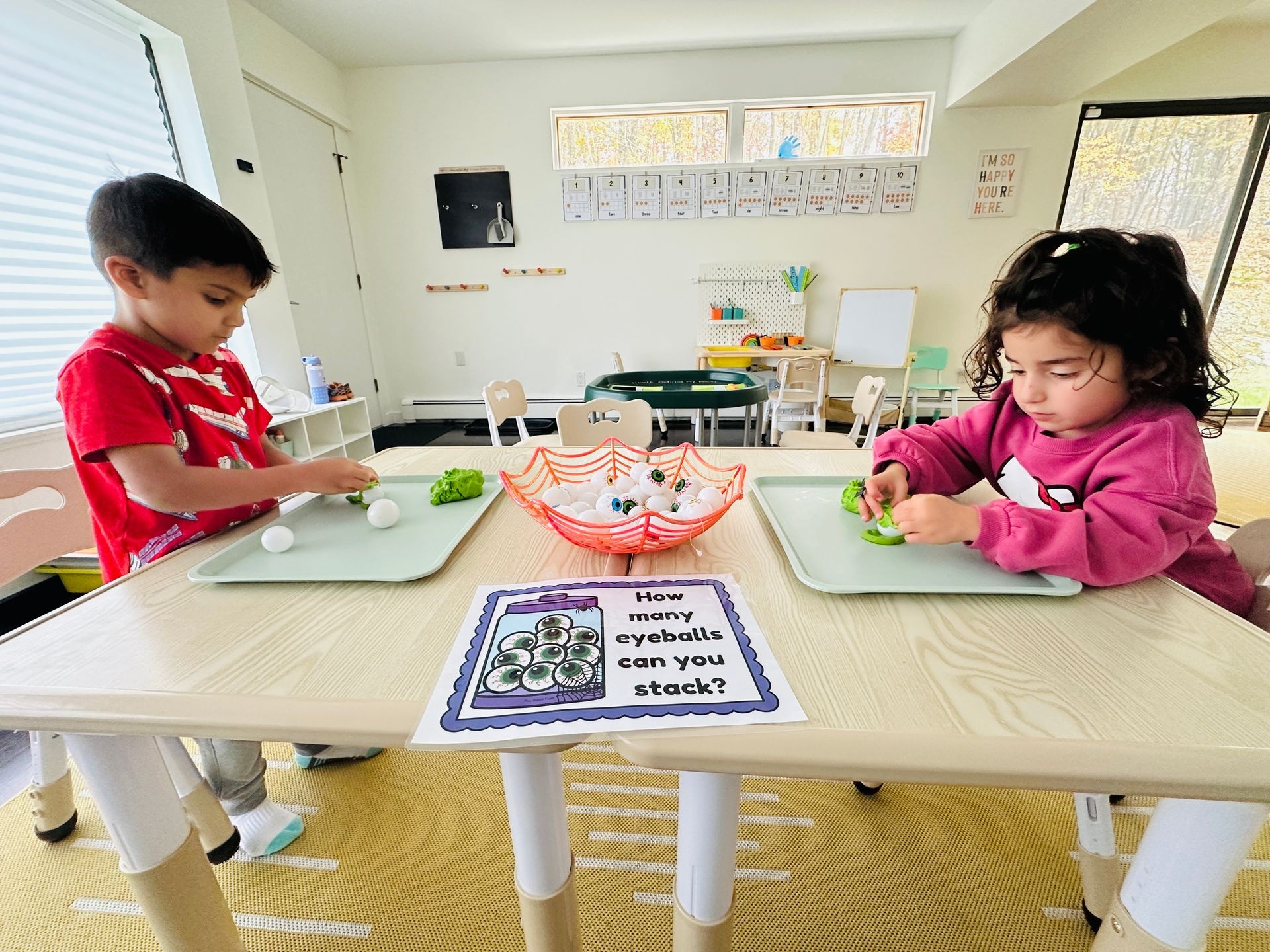 Two children sitting at a table stacking toy eyeballs on trays in a classroom setting.